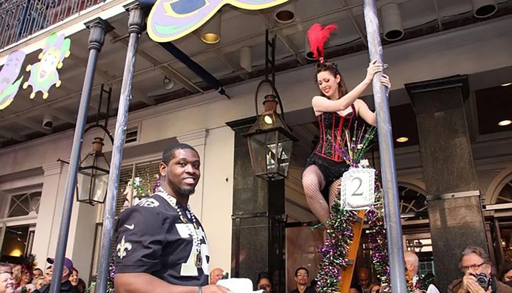 A performer in a burlesque-style outfit is on a pole above a smiling man in a football jersey amidst an audience suggesting a festive or celebratory event