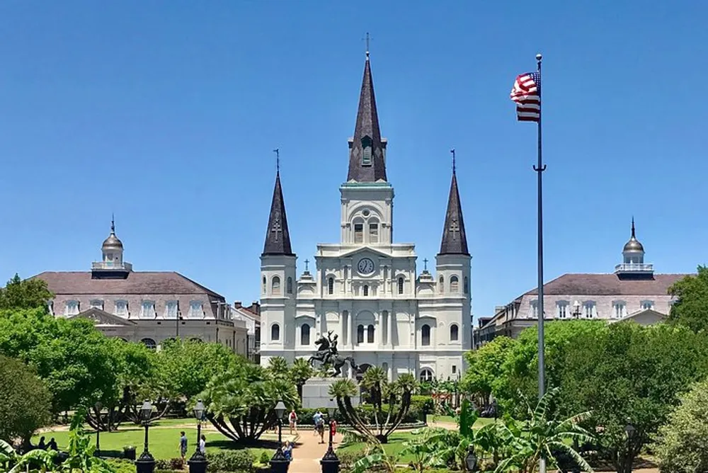 The image shows a historic cathedral with white faades and spires overlooking a green square with a statue under a clear blue sky with an American flag fluttering to the right