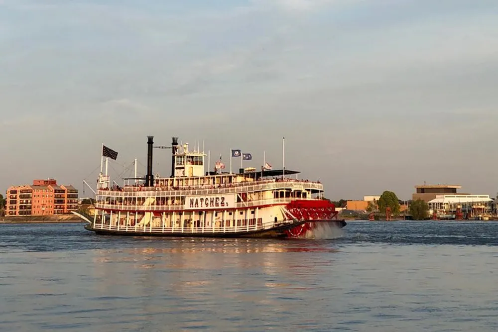 The image shows a traditional paddlewheel steamboat with the name Natchez on its side cruising on a large body of water during what appears to be late afternoon with buildings on the distant shoreline
