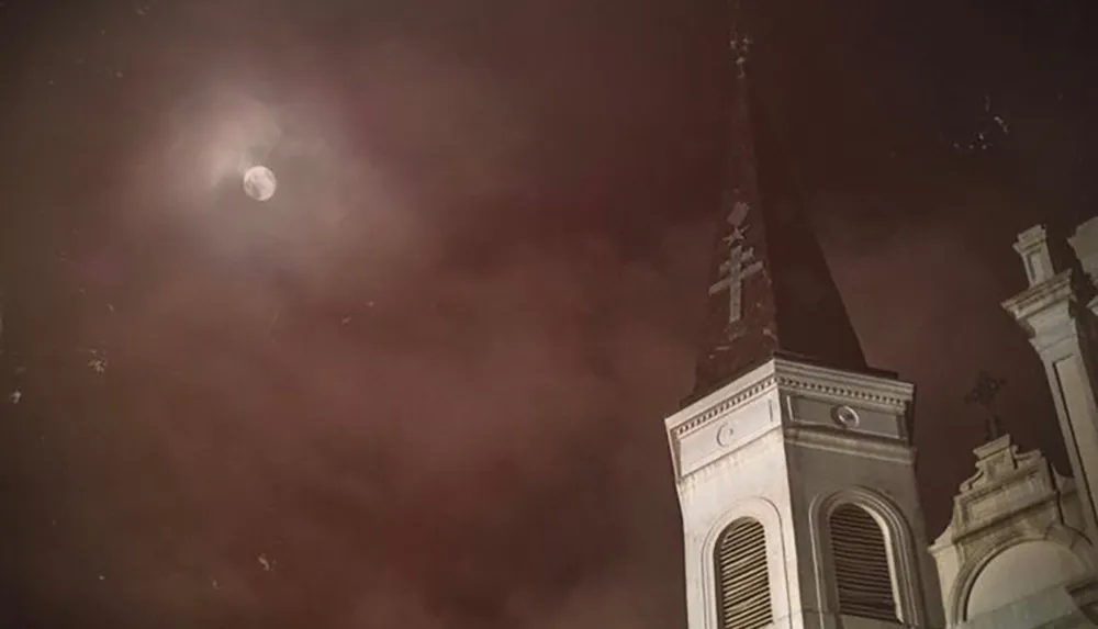 The image captures a moody nighttime scene with a full moon partially obscured by clouds looming over the silhouette of a church steeple