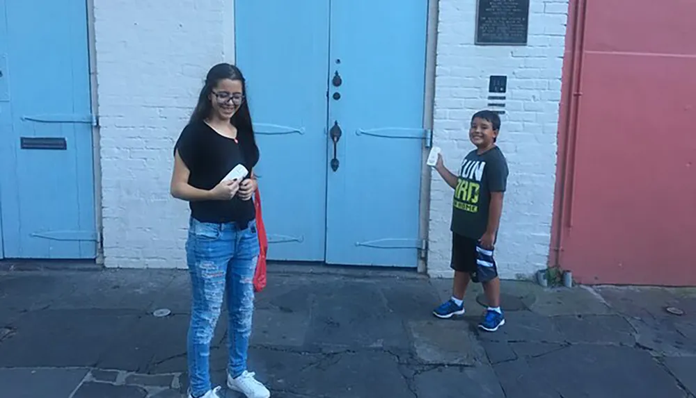 Two children are standing in front of a pair of bright blue doors set into a white brick wall each holding a mobile phone and smiling