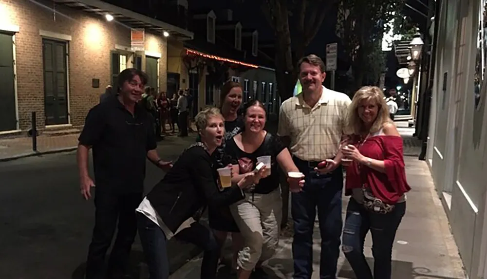 A group of people appear to be enjoying themselves on a lively street at night with some holding drinks and posing for the photo with expressions of laughter and smiles