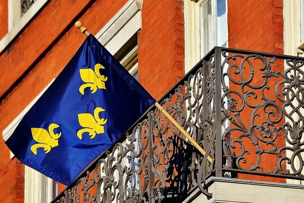 A blue flag with fleur-de-lis symbols is prominently displayed on a pole attached to an ornate iron balcony railing against a background of a red brick building