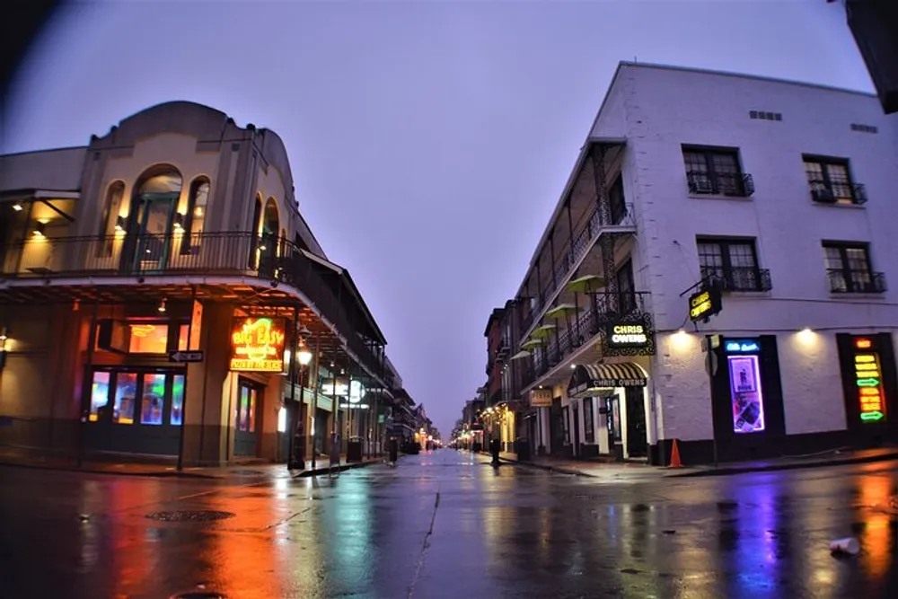 The image shows a wet street in an urban environment with historic architecture and neon signs during twilight or a rainy evening giving off a moody ambiance