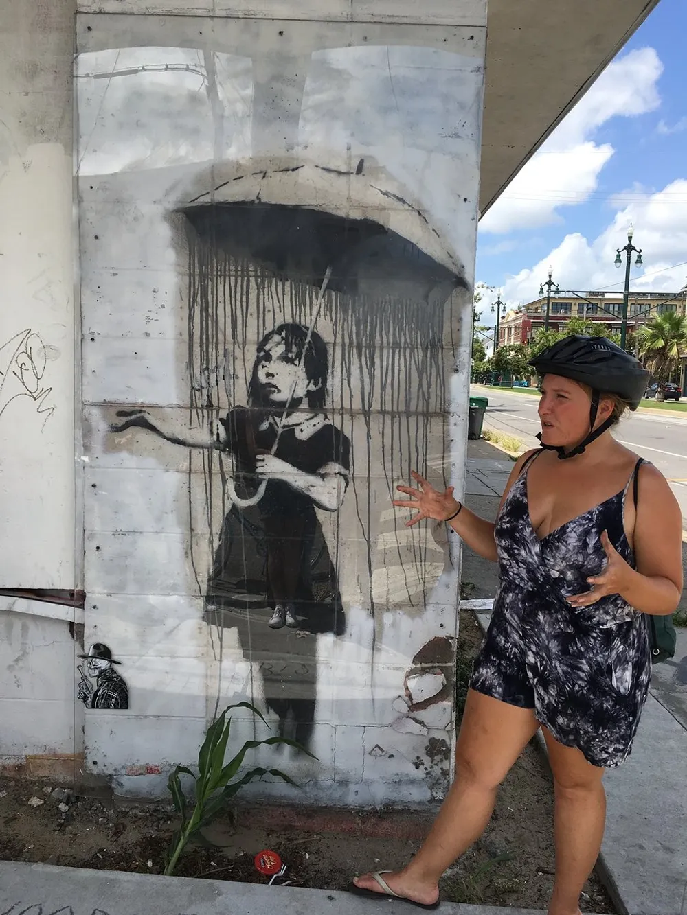 A person wearing a helmet is posing with an expressive gesture next to a street art piece depicting a child reaching out as if interacting with rain