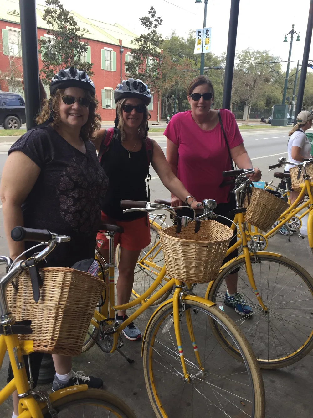 Three individuals wearing helmets stand smiling beside their yellow bicycles with wicker baskets apparently ready for a ride