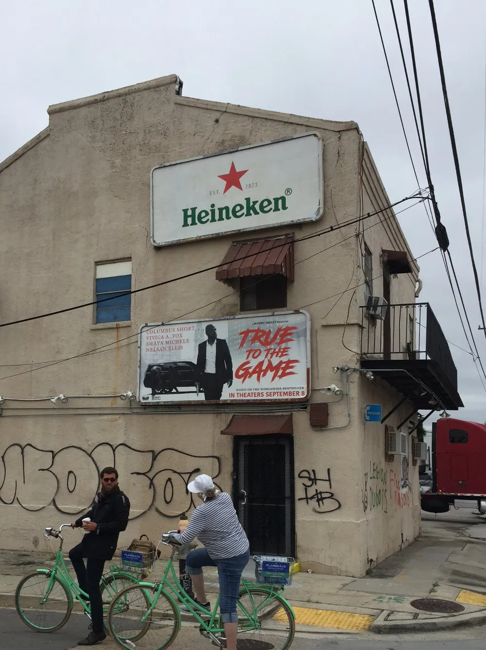 Two individuals on green bicycles are stopped at a corner in front of a building with a Heineken beer sign and a movie poster for True to the Game