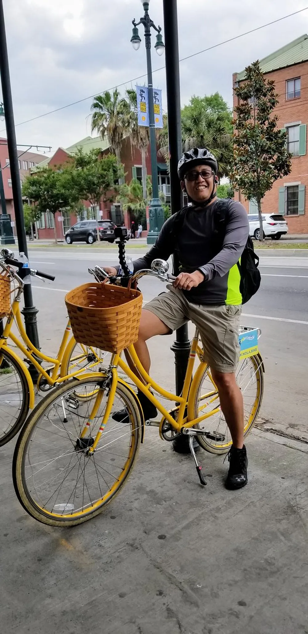 A person wearing a helmet and sunglasses is smiling at the camera while straddling a yellow rental bicycle with a basket in an urban setting near a tramway line