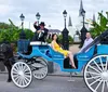 A man and a woman enjoy a carriage ride with the driver tipping his hat in front of an elegant gated area with historic architecture in the background