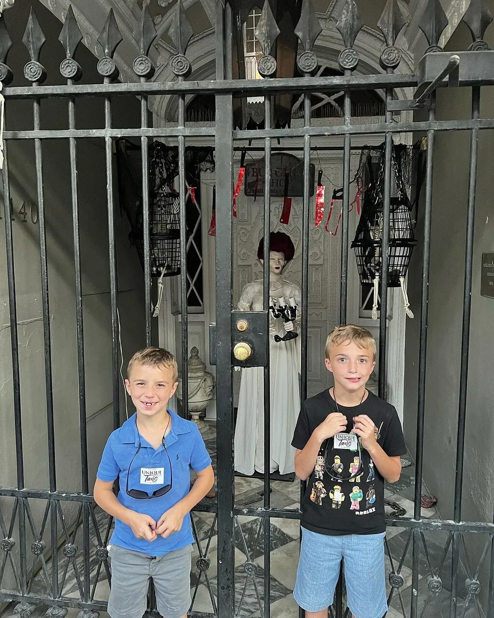 Two boys are smiling in front of a gate with an eccentrically dressed figure behind them creating a whimsical and curious scene