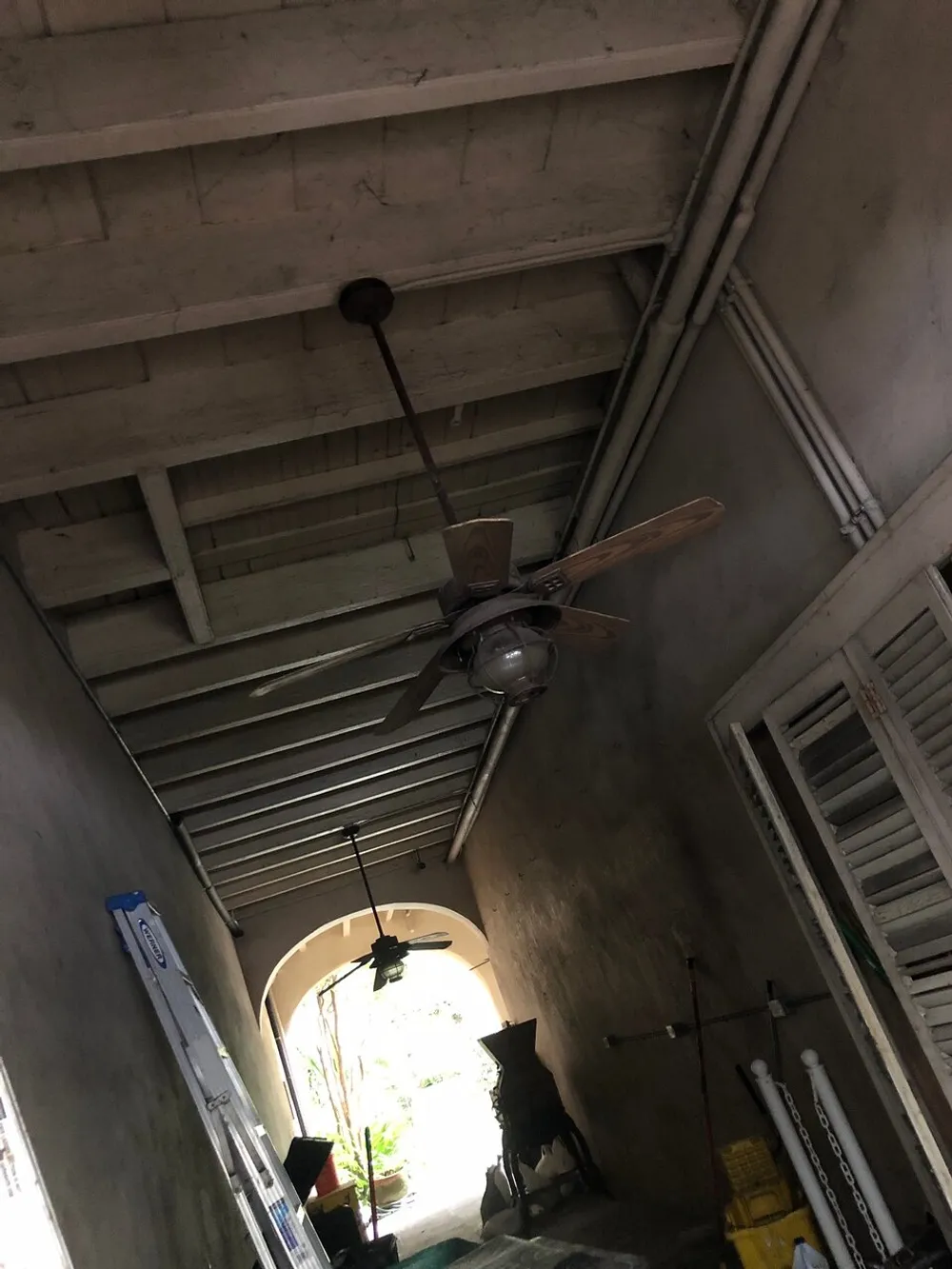 A ceiling fan is installed in a dimly lit narrow corridor with various items stored along the sides and daylight can be seen at the far end