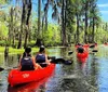 A woman is taking a selfie in the foreground while another person paddles a kayak in a swampy waterway behind her