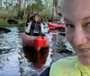 A woman is taking a selfie in the foreground while another person paddles a kayak in a swampy waterway behind her