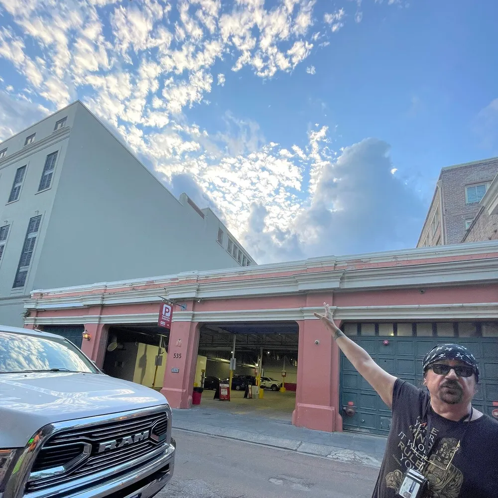 A man with sunglasses and a bandana is pointing upwards towards the beautiful sky next to an open-air parking structure with a pickup truck in the foreground