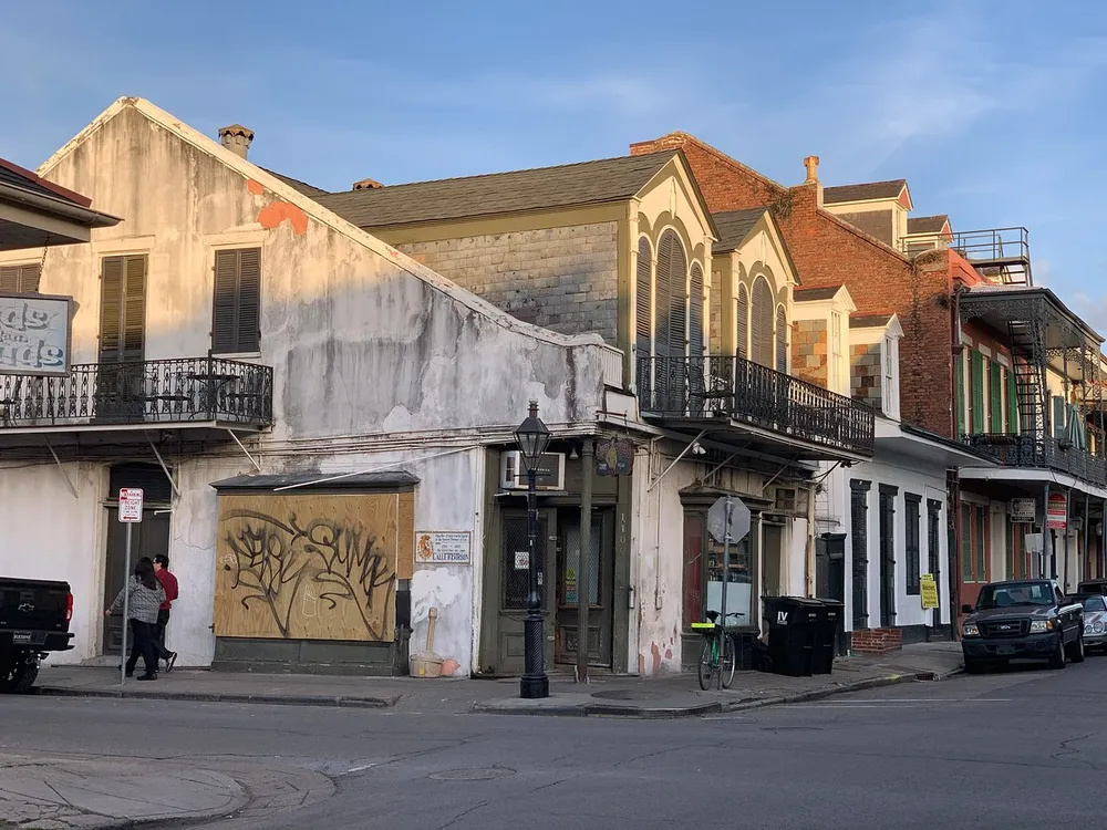 The image shows a street corner in what looks like the French Quarter of New Orleans with its historic buildings featuring balconies and a person walking past a boarded-up storefront