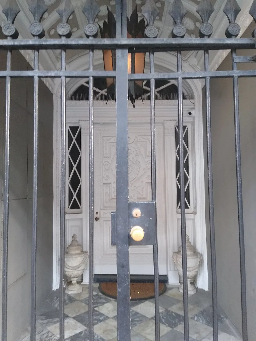 An ornate white door is visible behind a set of metal bars flanked by classical-style urns and complemented by a bell push button and intercom system