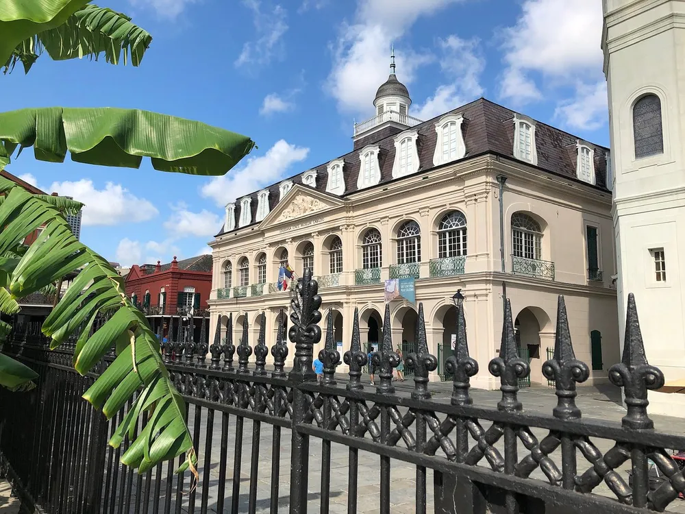 The image shows a historic building with classical architecture featuring a balcony and a mansard roof seen behind an ornate iron fence with tropical foliage in the foreground under a clear blue sky