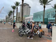 A group of people with bicycles, wearing helmets, are gathered on an urban waterfront promenade, possibly preparing for a ride or tour.