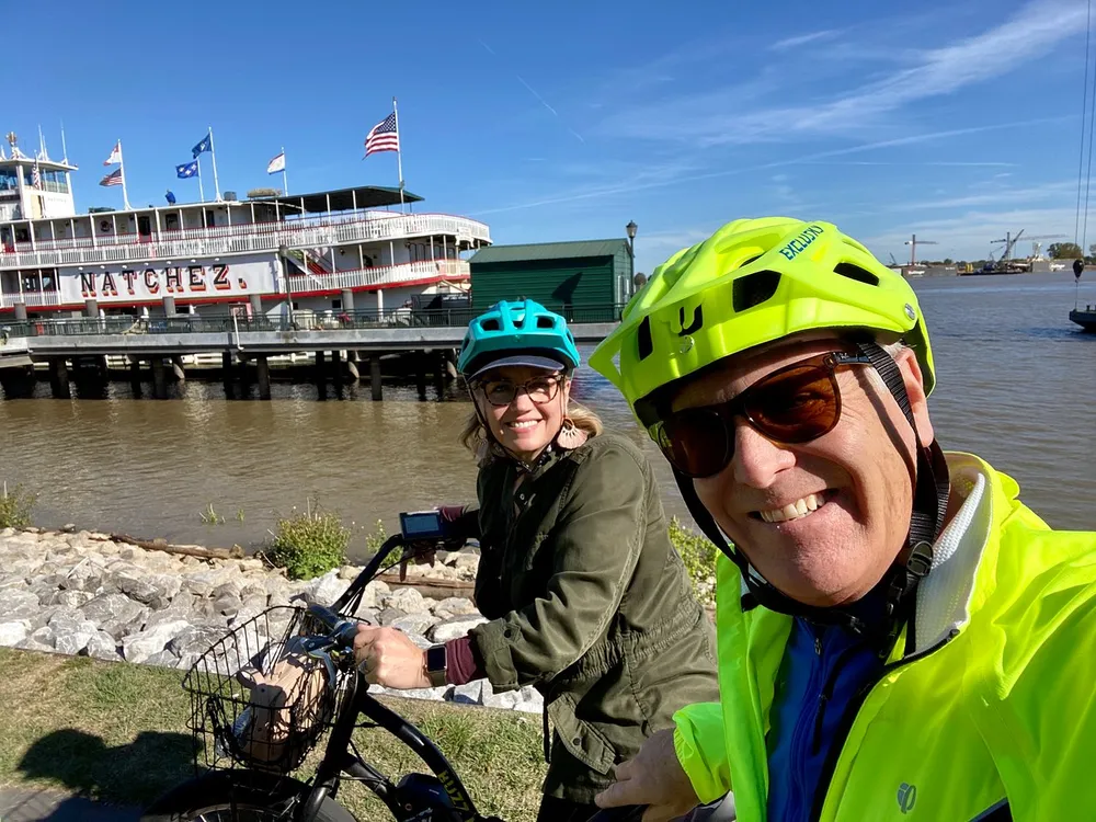 Two smiling people wearing bicycle helmets take a selfie with a river steamboat named Natchez in the background