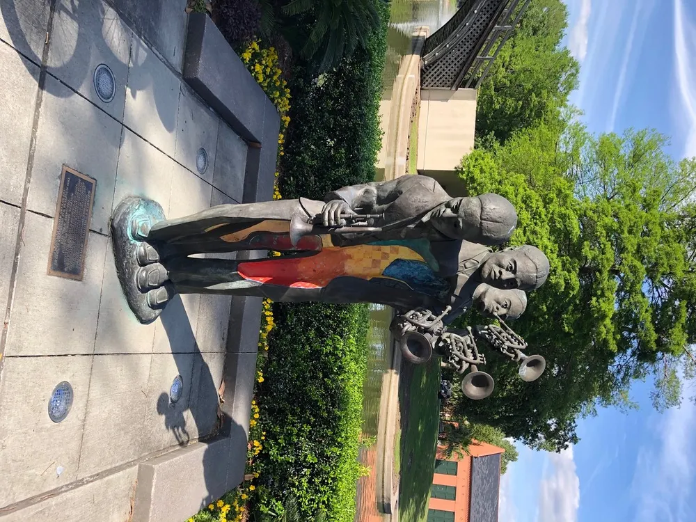 The image shows a colorful life-sized statue of three jazz musicians with their instruments in an outdoor setting bathed in sunlight and surrounded by greenery