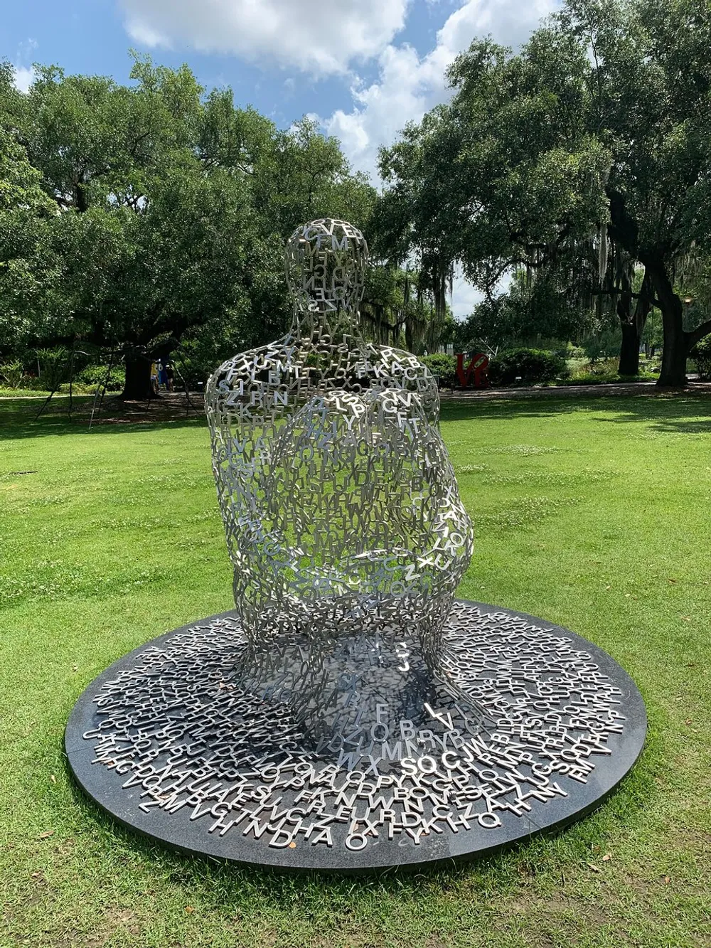 The image shows a unique outdoor sculpture of a seated human figure made of interconnected metal letters placed on a lush green lawn with trees in the background