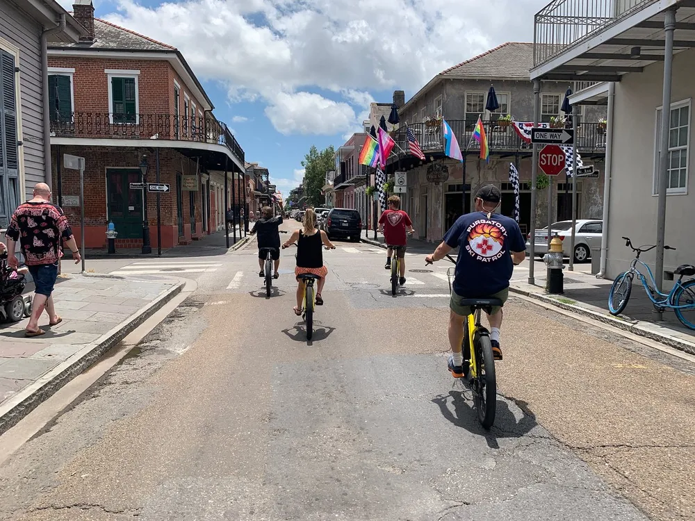 Three people are riding bicycles down a street in a bustling urban area characterized by historic architecture and vibrant flags