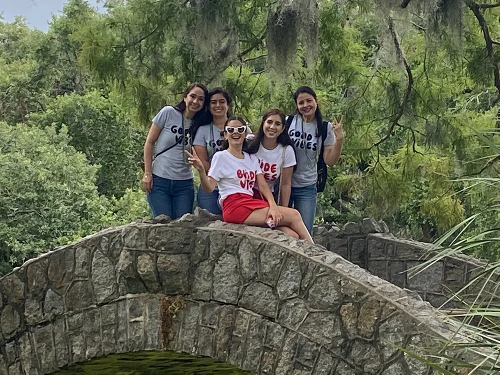 A group of five individuals is posing with peace signs and smiles atop a picturesque stone bridge surrounded by greenery