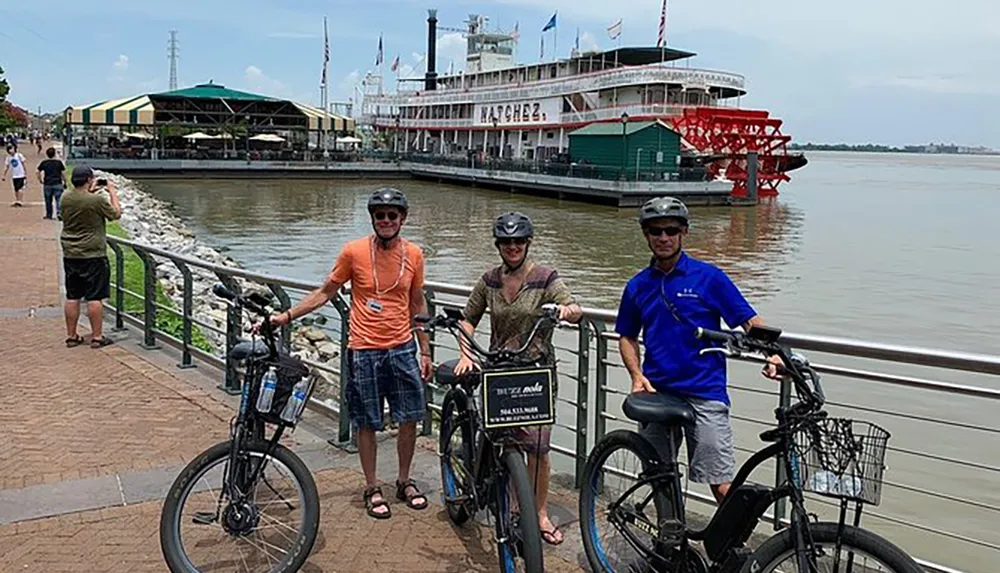 Three individuals are posing with their bicycles by a river railing in front of a paddle steamer