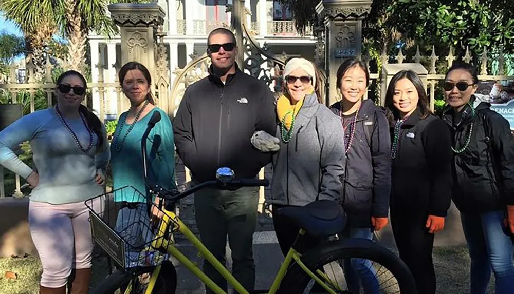 A group of smiling people some wearing Mardi Gras beads poses for a photo with a bicycle in front of an ornate gate and palm trees