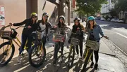 A group of five smiling people wearing helmets are posing with rental bicycles on a sunny city street.