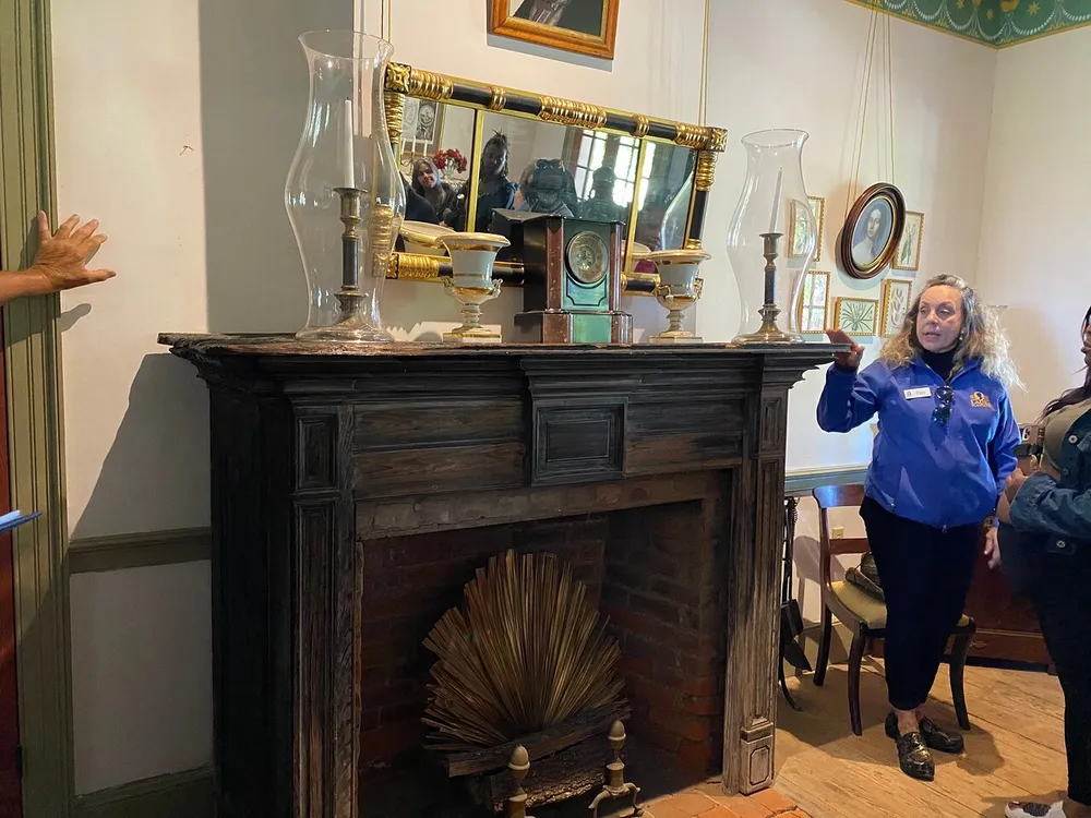 A tour guide is explaining features of a historical room with a decorative fireplace mantel objects and a mirror reflecting the scene