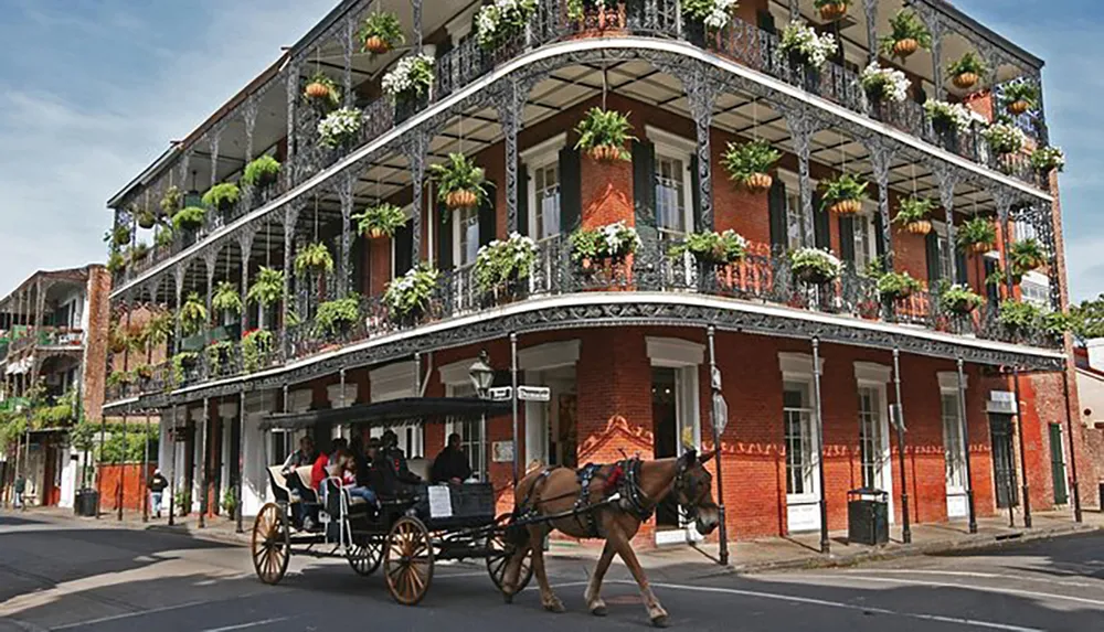 A horse-drawn carriage passes in front of a multi-storied building with ornate iron balconies adorned with hanging plants in a historic-looking urban setting