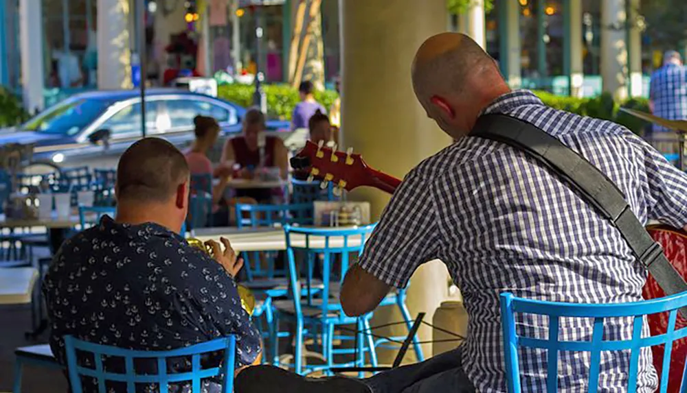 Two men sit at a sidewalk caf one playing guitar and the other looking at his phone with other patrons in the background and a car passing by