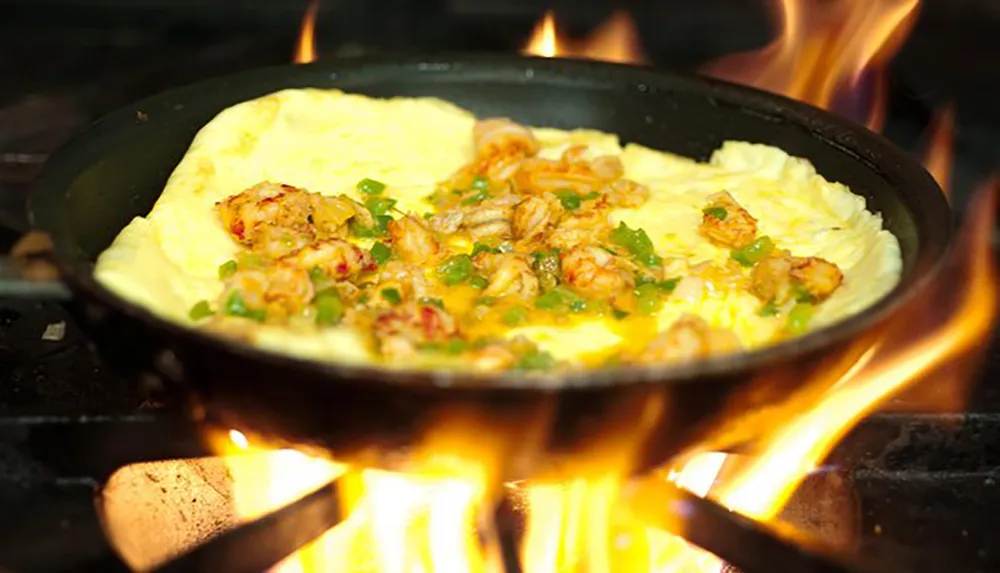 A skillet with an omelette garnished with green onions and pieces of shrimp is cooking over an open flame