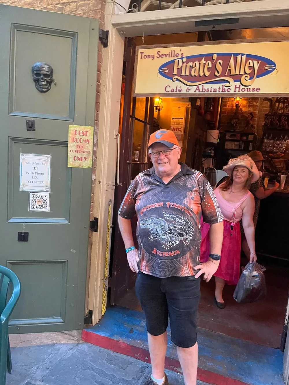 A man in a graphic shirt and a cap stands smiling in front of Tony Sevilles Pirates Alley Caf and Absinthe House with a woman peeking from behind and a decorative skull affixed to the green door on the left