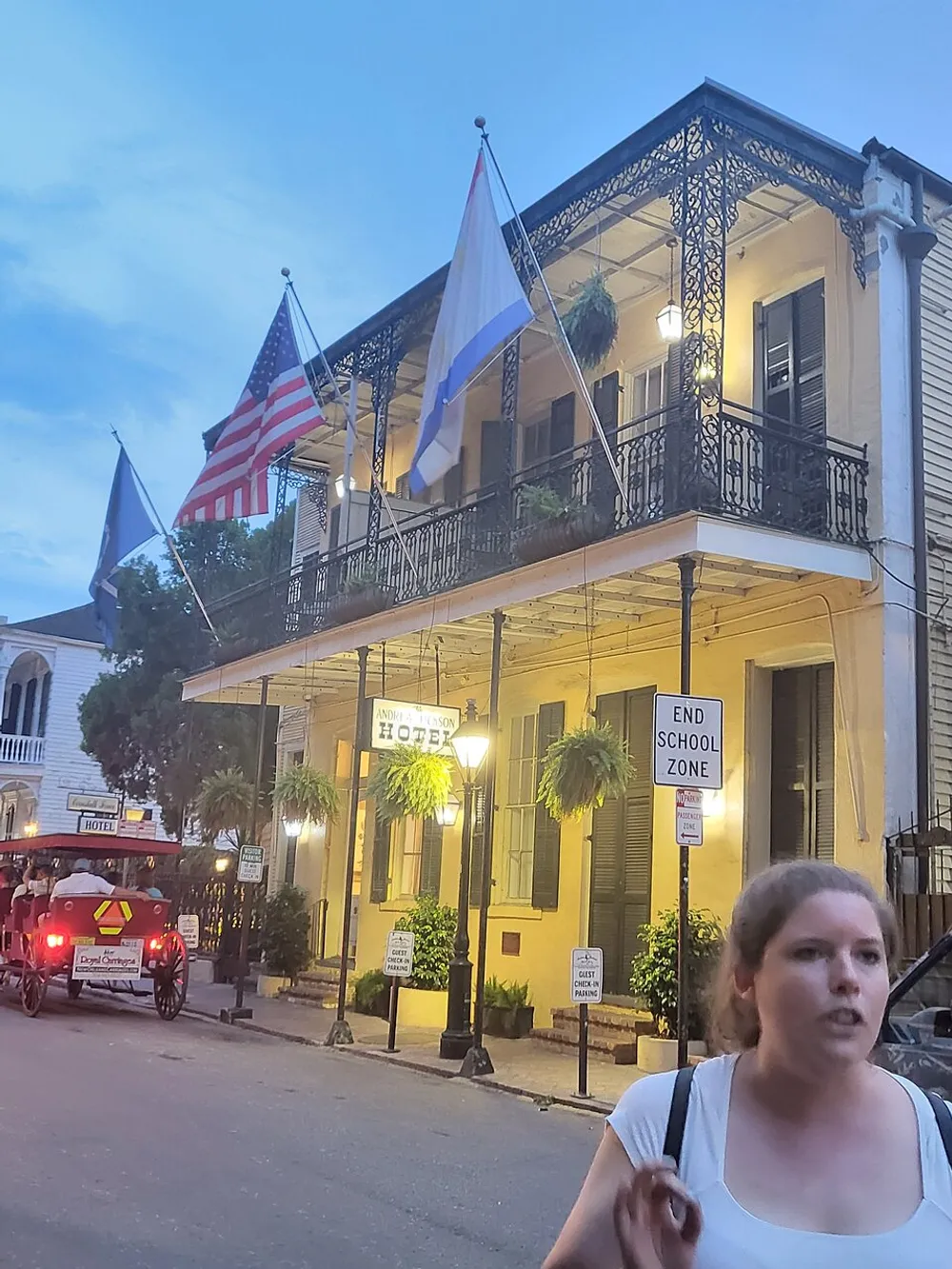 The image shows a two-story building with a balcony adorned with wrought-iron railings flying several flags with a sign indicating its a hotel a pedestrian on the street and a horse-drawn carriage passing by in the early evening