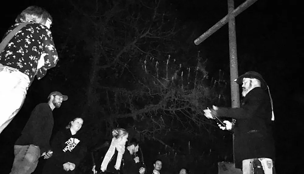 The black and white image features a group of people gathered at night in an outdoor setting with one individual elevated on a platform under a wooden structure pointing towards the audience which gives it a feel of an impromptu speech or performance