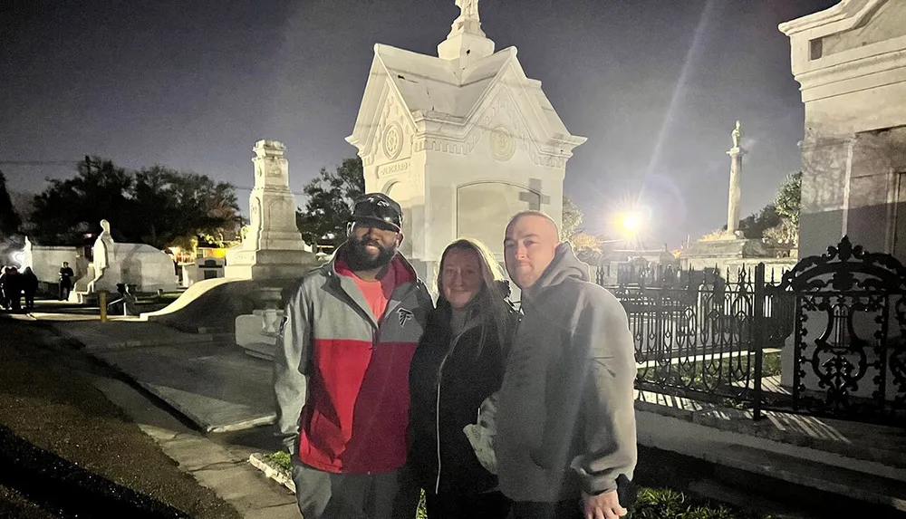 Three people are smiling for a photo in a well-lit cemetery at twilight