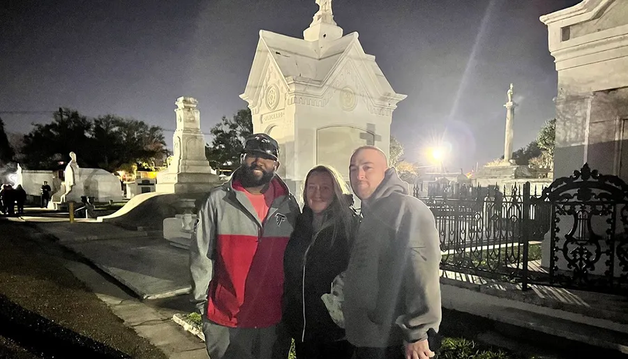 Three people are smiling for a photo in a well-lit cemetery at twilight.