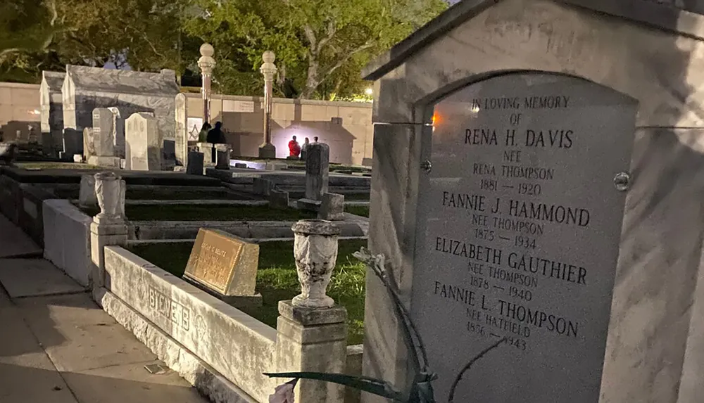 The image shows a nighttime view of a cemetery with various tombs and headstones some people in the distance and the illuminated grave marker of Rena H Davis in the foreground
