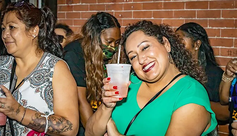A smiling woman in a green top holding a drink poses for the camera at what appears to be a social event