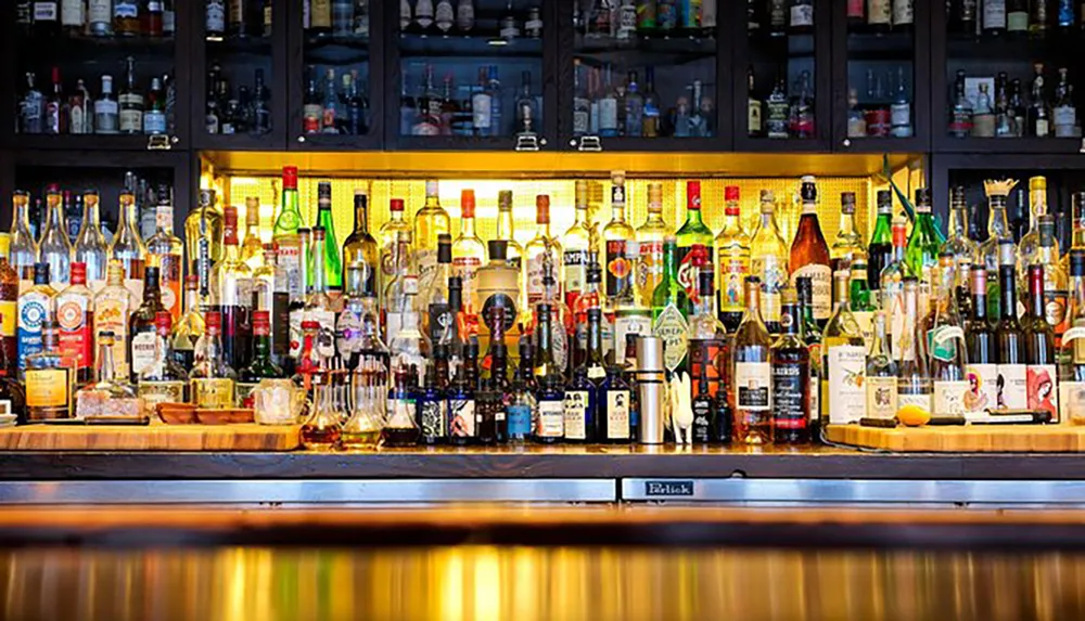 The image shows a well-stocked bar with various bottles of liquor displayed on shelves illuminated with warm backlighting