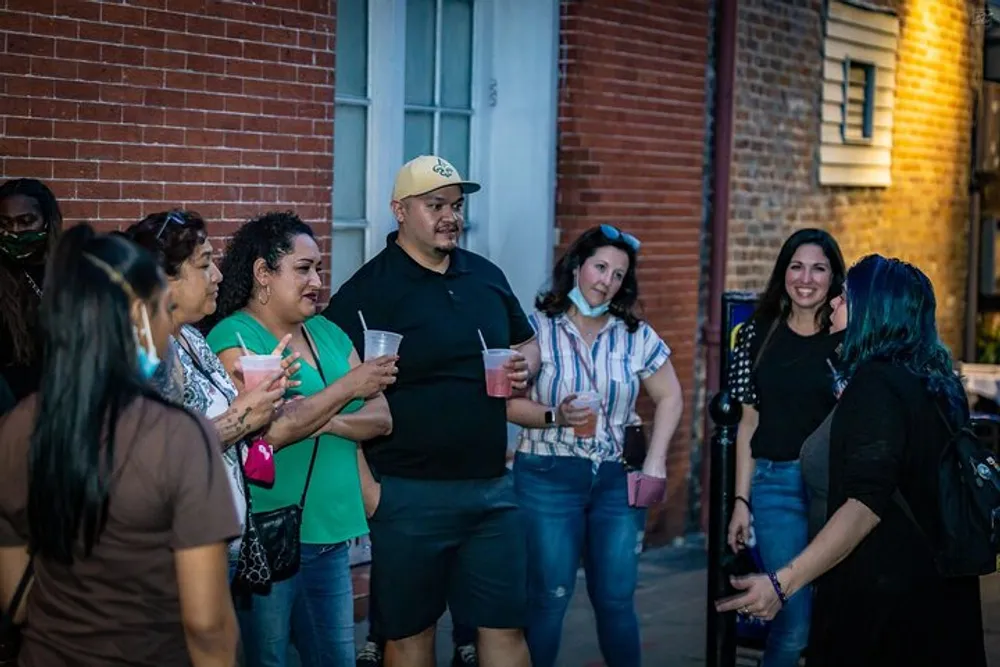 A group of people are casually socializing outdoors holding drinks and engaging in conversation with a brick building in the background and a warm evening atmosphere