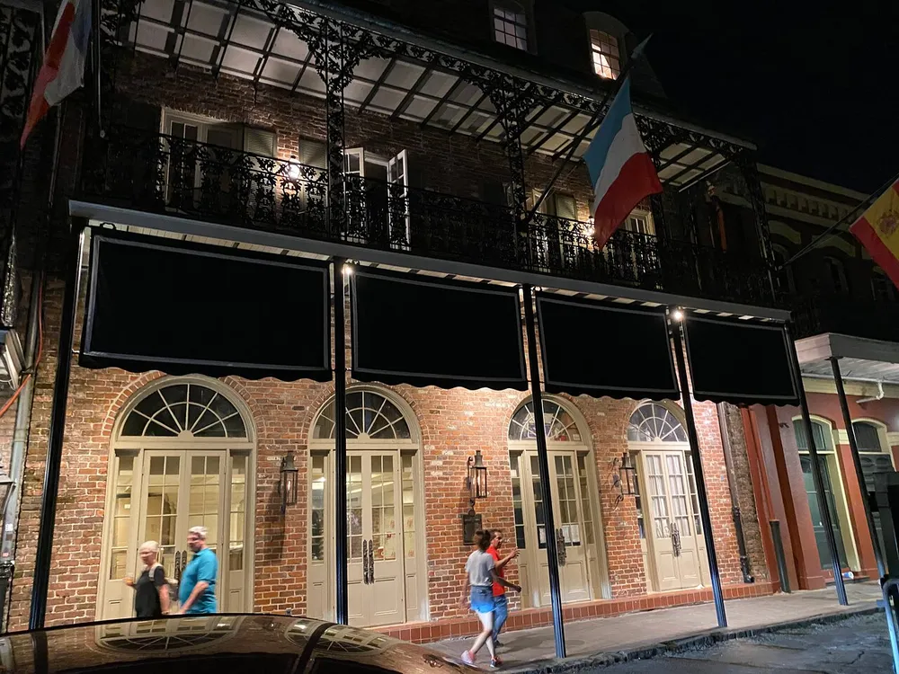A nighttime scene of a two-story brick building with arched windows and black awnings featuring pedestrians walking by and several flags hanging from the balcony