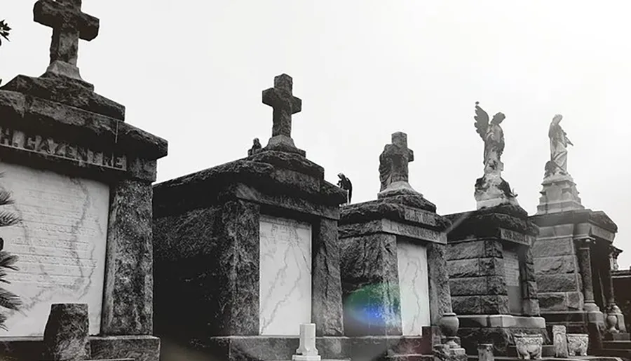 The image shows a black and white photograph of ornate cemetery monuments, topped with crosses and statues, conveying a somber and historical atmosphere.