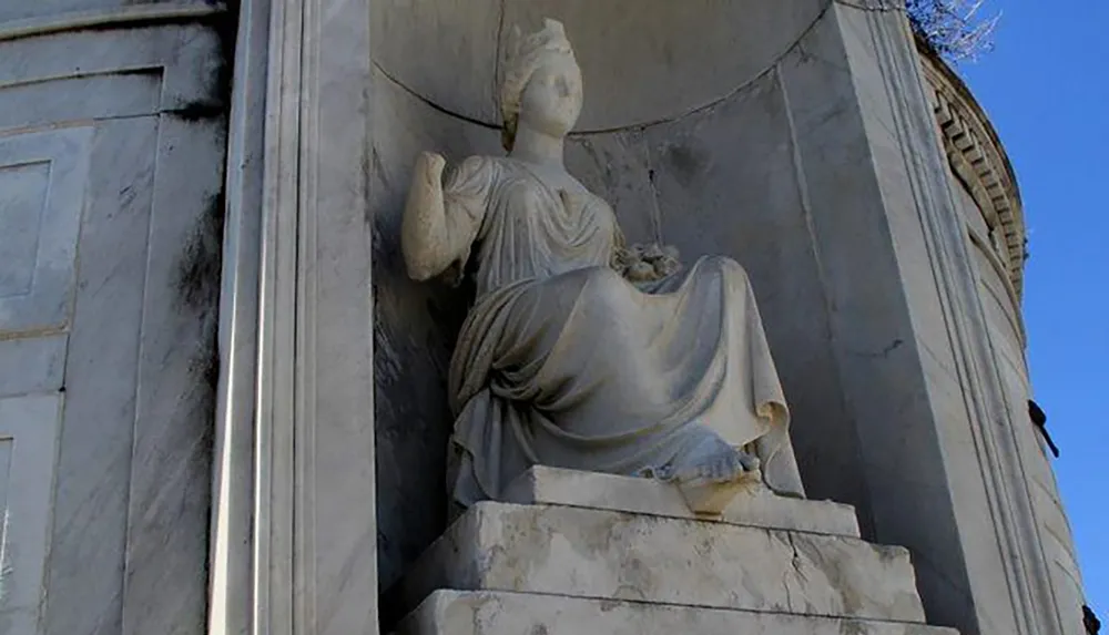 The photo depicts an ancient stone statue of a robed female figure seated on a throne-like chair with one arm raised set within a niche on a classical building
