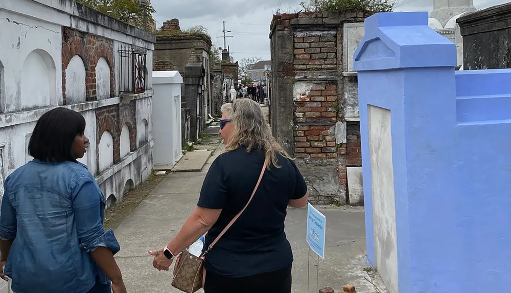 Two women are walking and conversing in a historic cemetery with above-ground tombs and weathered brickwork