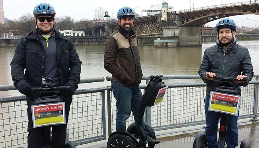 Three people are standing by a river each with a Segway and wearing helmets seemingly part of a Segway tour