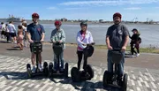 Four people are standing on Segways wearing helmets against a backdrop of a wide river and a crowd of pedestrians.