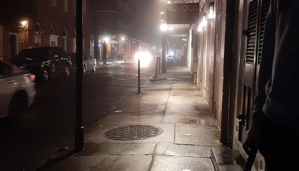 A dimly lit foggy urban street at night captured from a sidewalk perspective with cars parked and a glowing street lamp in the distance